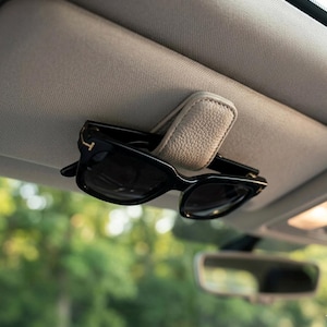 Op de afbeelding: Een beige leren zonnebrilhouder bevestigd aan een auto zonneklep, met een zwarte zonnebril. De achtergrond is wazig en toont een auto-interieur en groen. Handig voor het opbergen van brillen.
