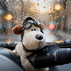 Op de afbeelding: Een pluchen hond met een leren pilotenhelm en bril in een auto. De hond houdt een zwarte auto-bedieningshendel vast. Regendruppels zijn zichtbaar op de voorruit, met vage stadslichten op de achtergrond.