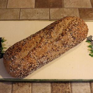 May include: A loaf of seeded bread rests on a rectangular, off-white serving board. The bread is brown with a mix of seeds. The board has a green border and decorative black and green accents.