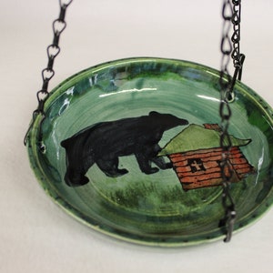 May include: A hanging, green ceramic bird feeder with a black chain. The feeder features a hand-painted design of a black bear and a small cabin. The interior of the feeder is a lighter green.