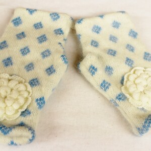 May include: A pair of ivory-colored mittens with a blue diamond pattern and a cream-colored flower applique. The cuffs and edges are trimmed with blue. These mittens are designed for warmth and style.