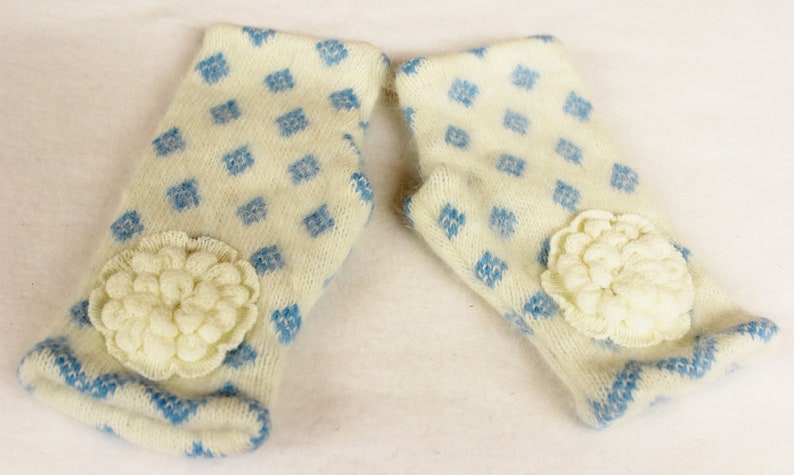 May include: A pair of ivory-colored mittens with a blue diamond pattern and a cream-colored flower applique. The cuffs and edges are trimmed with blue. These mittens are designed for warmth and style.