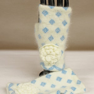 May include: Cream-colored fingerless gloves with a blue diamond pattern and a cream-colored flower accent. The gloves are made of a soft, fuzzy material. The gloves are displayed on a black hand.