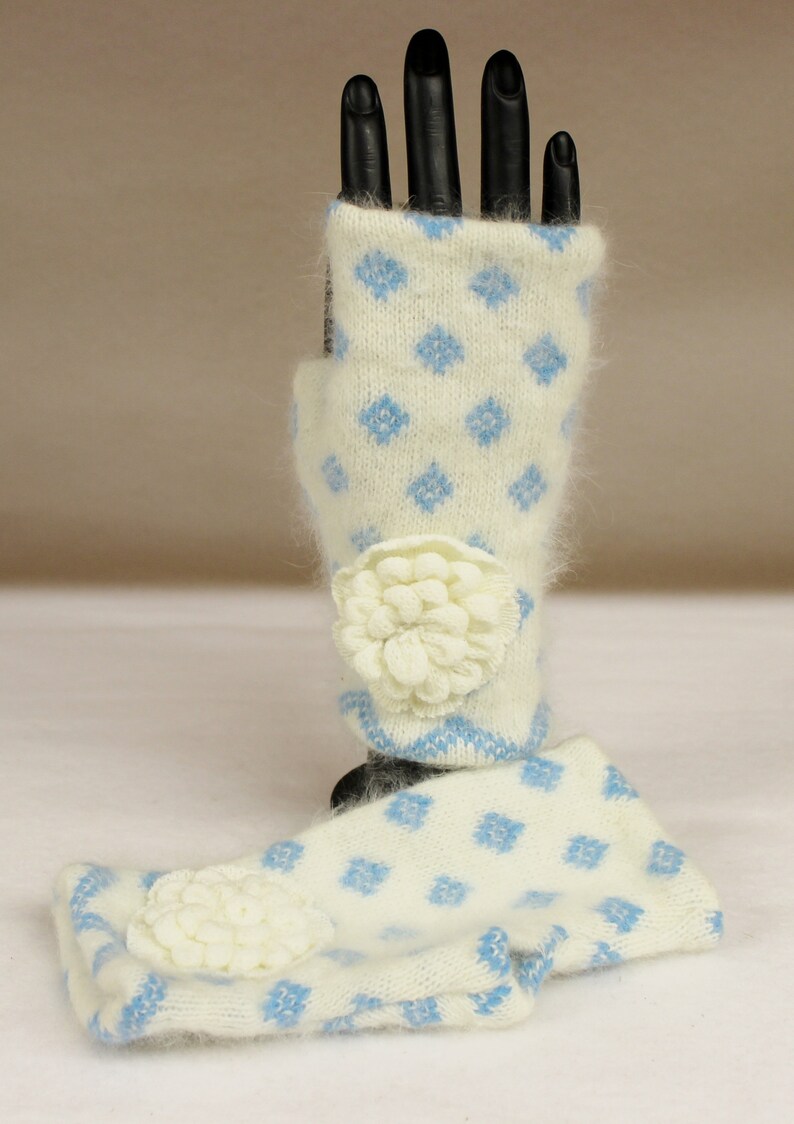 May include: Cream-colored fingerless gloves with a blue diamond pattern and a cream-colored flower accent. The gloves are made of a soft, fuzzy material. The gloves are displayed on a black hand.