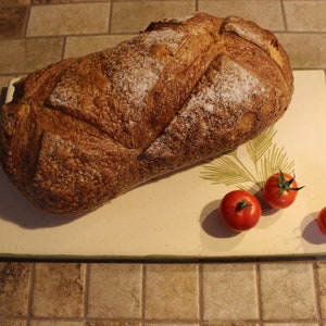 May include: A loaf of crusty bread with a scored top rests on a rectangular cutting board with a green border and a fern design. Three red tomatoes with green stems are arranged near the bread.