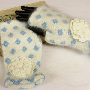 May include: A pair of ivory-colored fingerless gloves with a light blue diamond pattern. Each glove features a cream-colored, flower-shaped embellishment. The cuffs and edges are trimmed with light blue. The gloves are made of a soft, fuzzy material.