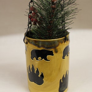 May include: A hanging ceramic planter with a glossy, golden-brown finish. The planter features black bear silhouettes and paw prints. It is suspended by a black chain and filled with faux pine branches and red berries, creating a rustic, nature-inspired aesthetic.