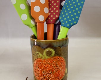 May include: A collection of colorful silicone spatulas with white polka dots, in green, orange, red, and blue, are displayed in a ceramic pumpkin-themed holder. The holder is brown with an embossed orange pumpkin design.