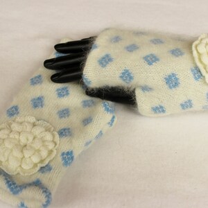 May include: A pair of ivory-colored fingerless gloves with a blue diamond pattern. Each glove features a large, cream-colored, flower-shaped embellishment. The cuffs are trimmed with blue. The gloves are made of a soft, fuzzy material.