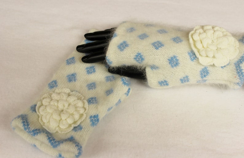 May include: A pair of ivory-colored fingerless gloves with a blue diamond pattern. Each glove features a large, cream-colored, flower-shaped embellishment. The cuffs are trimmed with blue. The gloves are made of a soft, fuzzy material.