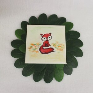 May include: A small square canvas painting featuring a red fox illustration. The fox is depicted in a seated position with a white belly and black accents. The painting is surrounded by green leaves, creating a decorative border.