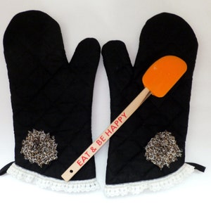 May include: A pair of black quilted oven mitts with a white lace trim and a decorative floral accent. An orange spatula with a wooden handle that reads "EAT & BE HAPPY" is placed between the mitts.