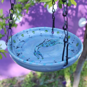 May include: A hanging, hand-painted bird bath with a light blue base and a decorative fish design. The bird bath is suspended by black chains and features green and turquoise accents. A purple background is visible.