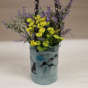 May include: A hanging ceramic planter with a mountain and wolf design. The planter is filled with yellow and purple flowers. The planter is a light blue color and is suspended by a black chain.