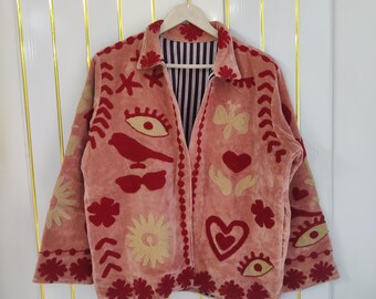 May include: A pink jacket with embroidered details. The jacket features red and gold-colored embroidered designs of eyes, birds, hearts, and flowers. The jacket has a collared neckline and a striped interior lining.