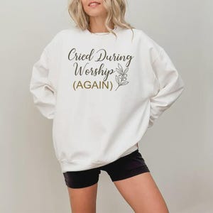 Puede incluir: Sudadera blanca con el texto "Cried During Worship (AGAIN)" en fuente cursiva, adornada con una ilustración botánica. La sudadera se combina con pantalones cortos negros.