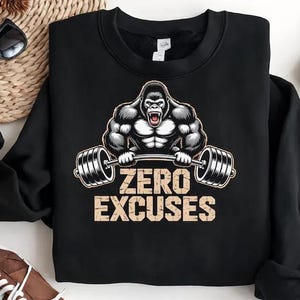Puede incluir: Sudadera negra con un gorila musculoso levantando una barra. El diseño incluye el texto "ZERO EXCUSES" en una fuente desgastada. La imagen también muestra gafas de sol, una esterilla tejida, vaqueros y zapatillas marrones.