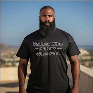 May include: Black t-shirt with the text "Protect What Matters - Work Safe." The shirt is short-sleeved and has a crew neck. The image shows a person wearing the shirt against a blurred outdoor background.