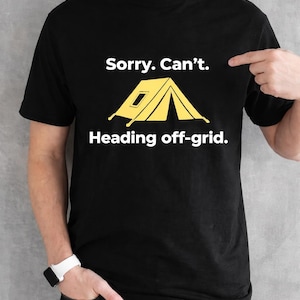 May include: Black t-shirt with the text "Sorry. Can't. Heading off-grid." and a yellow tent graphic. The shirt is being worn by a person. The person is wearing a white watch and dark jeans.