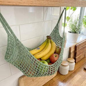 May include: A light green, crocheted fruit hammock filled with yellow bananas and red and green apples. The hammock hangs from a metal hook on a white tiled wall. The kitchen setting includes wooden cutting boards and jars.