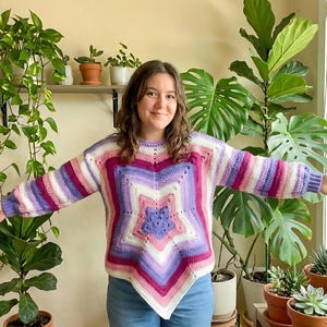 Peut inclure: Un pull crocheté à la main avec un motif d'étoile dans les tons de rose, de violet et de blanc. Le pull a des manches longues avec des rayures et un col rond. Le vêtement est présenté dans une pièce avec des plantes en pot.