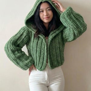 以下が含まれることがあります： 緑色の、リブ編み模様のフード付きかぎ針編みジャケット。長袖でゆったりとしたフィット感。フロントオープンデザインとフードがカジュアルで快適なスタイルを演出。ジャケットは無地のミディアムグリーンです。