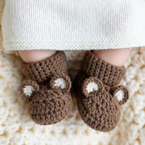 May include: Brown crochet baby booties with bear ears and white inner ear detail. The booties are on a cream-colored crocheted blanket. The baby is wearing a white knitted outfit.