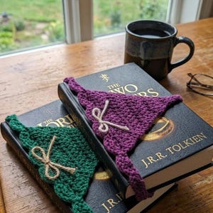 Puede incluir: Dos marcapáginas de ganchillo, uno verde y otro morado, descansan sobre un libro. Cada marcapáginas tiene un lazo de cuerda. Una taza de café y unas gafas también son visibles. El libro se titula "The Two Towers" de J.R.R. Tolkien.
