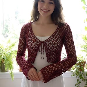 Puede incluir: Un bolero de ganchillo burdeos con mangas largas y cierre de lazo. El diseño calado crea un patrón geométrico. El bolero se lleva sobre un vestido blanco. El fondo incluye plantas verdes y una ventana.