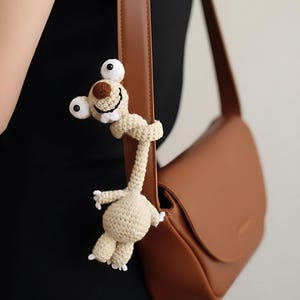 Pode incluir: Um amuleto de bicho-preguiça bege de crochê com olhos brancos grandes, um nariz castanho e uma boca sorridente. A preguiça está pendurada em uma alça de bolsa marrom. A bolsa é de cor castanho claro.