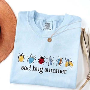 May include: Pale blue t-shirt featuring embroidered insects in various colours and the phrase "sad bug summer". A gold necklace with a pendant is draped over the shirt. The shirt is folded on a white surface.