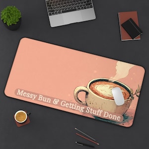 May include: A rectangular desk mat with a peach-colored background featuring an illustration of a coffee mug and the text "Messy Bun & Getting Stuff Done". The mat has a white computer mouse resting on the mug illustration.