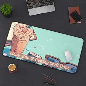 May include: A rectangular desk mat featuring an illustrated light blue background with a large iced coffee, a smartphone, and sunglasses. The text "Messy Bun & Getting Stuff Done" is printed in white across the center of the mat over the items.