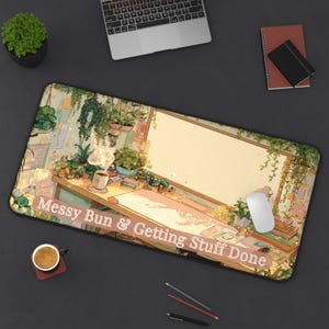 May include: A rectangular desk mat featuring an illustrated workspace scene with hanging plants, a desk, books, and a steaming mug. The text "Messy Bun & Getting Stuff Done" is printed in white across the bottom section of the mat's surface.
