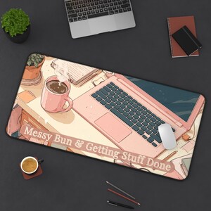 May include: A rectangular desk mat featuring an anime-style illustration of a workspace with a pink laptop, a pink mug, and a notebook. The text "Messy Bun & Getting Stuff Done" is printed in white across the bottom. The design includes a soft color palette of pinks, creams, and browns, depicting a cozy office scene with a computer mouse and stationery items.