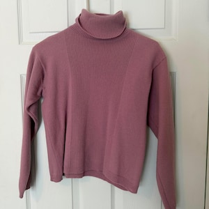 May include: A mauve-colored turtleneck sweater hanging on a white hanger. The sweater has a ribbed design on the front and a smooth texture on the sleeves. The turtleneck is folded over. The sweater is made of a soft material.
