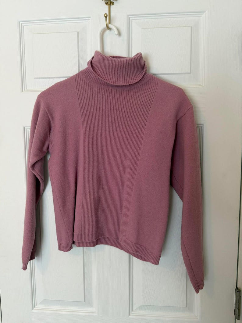 May include: A mauve-colored turtleneck sweater hanging on a white hanger. The sweater has a ribbed design on the front and a smooth texture on the sleeves. The turtleneck is folded over. The sweater is made of a soft material.