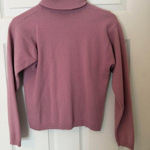 May include: A mauve-colored turtleneck sweater with long sleeves. The sweater is hanging on a white hanger against a white door. The turtleneck is ribbed, and the sleeves and hem have a ribbed finish.