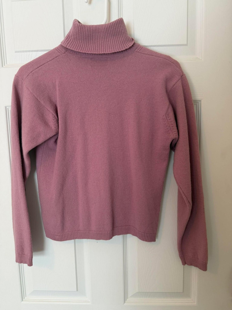 May include: A mauve-colored turtleneck sweater with long sleeves. The sweater is hanging on a white hanger against a white door. The turtleneck is ribbed, and the sleeves and hem have a ribbed finish.