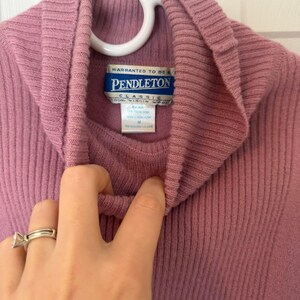 May include: A mauve-colored turtleneck sweater with a ribbed texture, hanging on a white hanger. The label reads "Pendleton Classic 100% Virgin Wool". The sweater is a medium size.