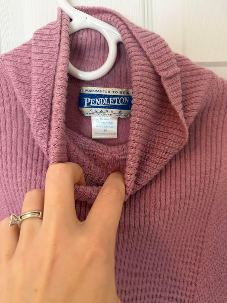May include: A mauve-colored turtleneck sweater with a ribbed texture, hanging on a white hanger. The label reads "Pendleton Classic 100% Virgin Wool". The sweater is a medium size.