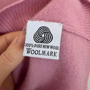 May include: Close-up of a pink knit garment with a white label. The label features a black and white circular design and the text "100% PURE NEW WOOL WOOLMARK". The garment is made of a soft, textured material.
