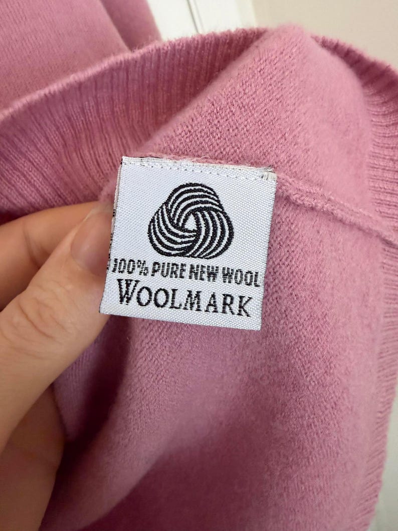 May include: Close-up of a pink knit garment with a white label. The label features a black and white circular design and the text "100% PURE NEW WOOL WOOLMARK". The garment is made of a soft, textured material.