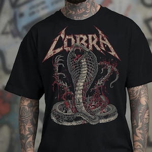 May include: Black t-shirt featuring a detailed graphic of a cobra with its mouth open, and the word "COBRA" in a stylized font. The cobra design is in shades of gray and red, with a grunge-style background.