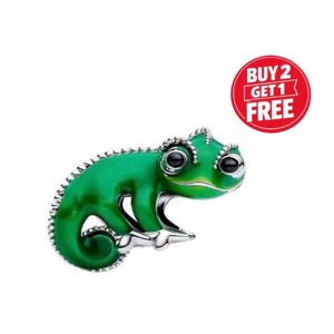 May include: A green chameleon-shaped pendant with black eyes and silver accents. The pendant has a glossy finish and is set against a white background. A red circle with the text "BUY 2 GET 1 FREE" is in the upper right corner.