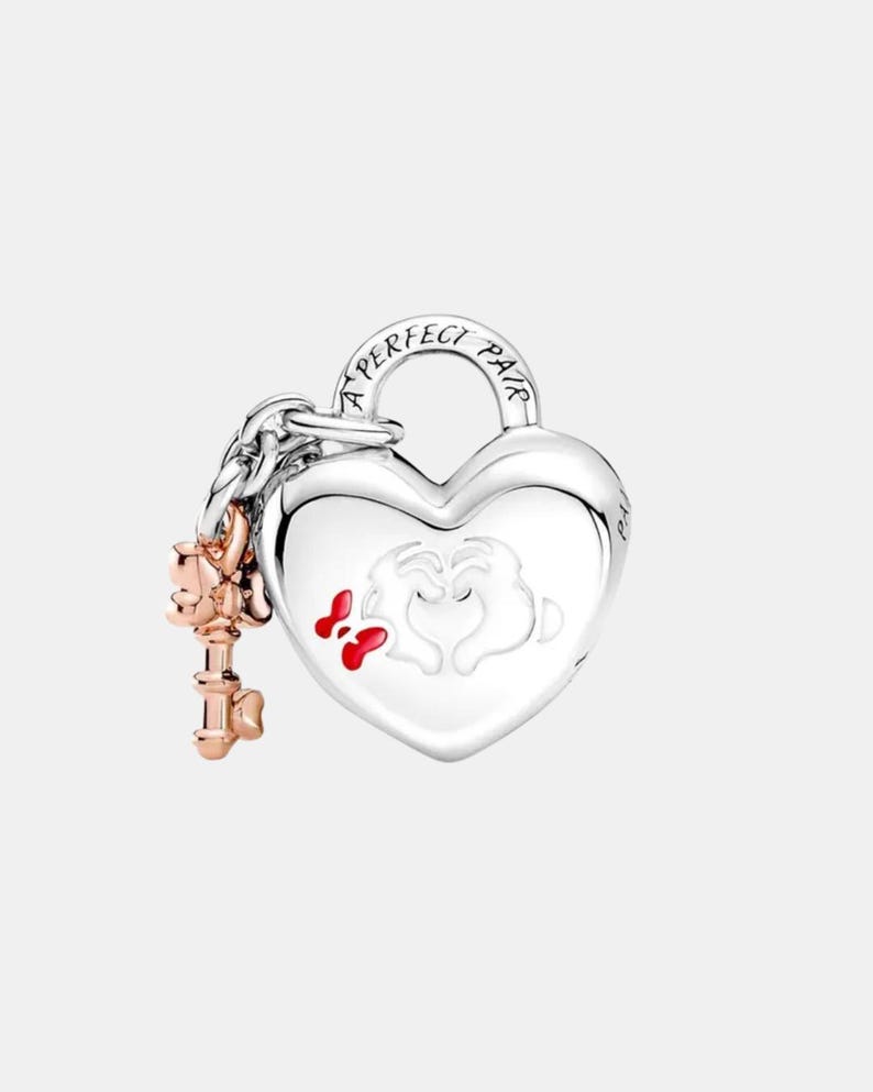 May include: A silver heart-shaped charm with the words "A PERFECT PAIR" engraved on the top. The charm features a design of two hands forming a heart with red accents. A rose gold key charm hangs from a chain attached to the heart.