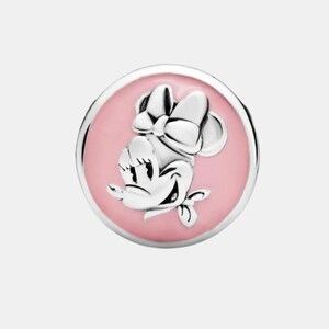 May include: A silver-colored charm featuring Minnie Mouse with a pink enamel background. The charm has a round shape with a raised Minnie Mouse design, including her signature bow. The charm is likely made of sterling silver.