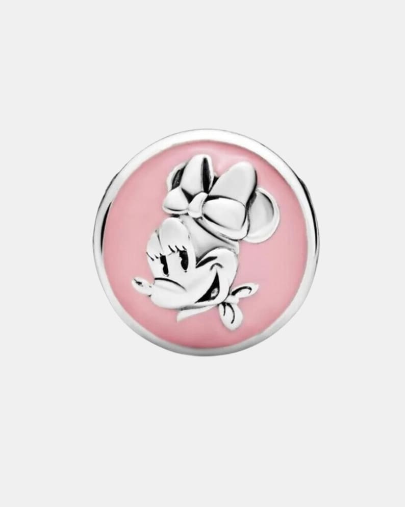 May include: A silver-colored charm featuring Minnie Mouse with a pink enamel background. The charm has a round shape with a raised Minnie Mouse design, including her signature bow. The charm is likely made of sterling silver.