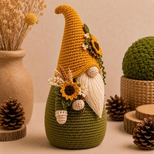 May include: A crochet gnome figurine featuring a tall mustard yellow pointed hat adorned with a sunflower and small white flowers. The gnome has a cream-colored yarn beard, a green body, and a decorative arrangement of sunflowers and wheat.