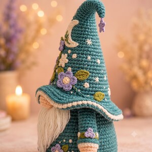 May include: A teal crochet gnome figurine featuring a tall, pointed hat decorated with a white crescent moon, small white embroidered stars, and several purple and white crocheted flowers with green leaves. The gnome has a long, white yarn beard.